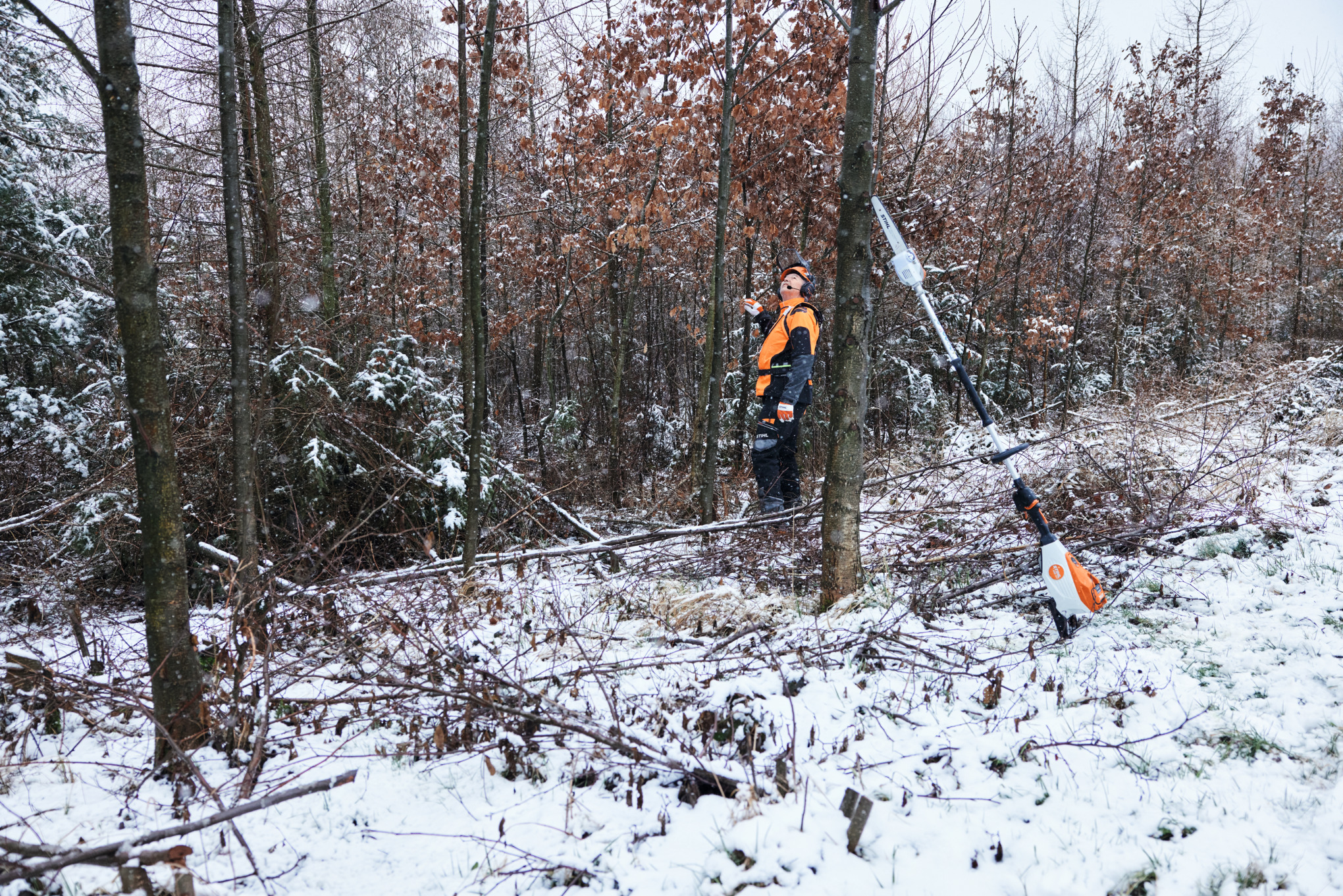 Een man werkt in veiligheidsuitrusting in een besneeuwd bos met op de voorgrond een STIHL HTA 150 accuhoogsnoeier die tegen een boomstam staat