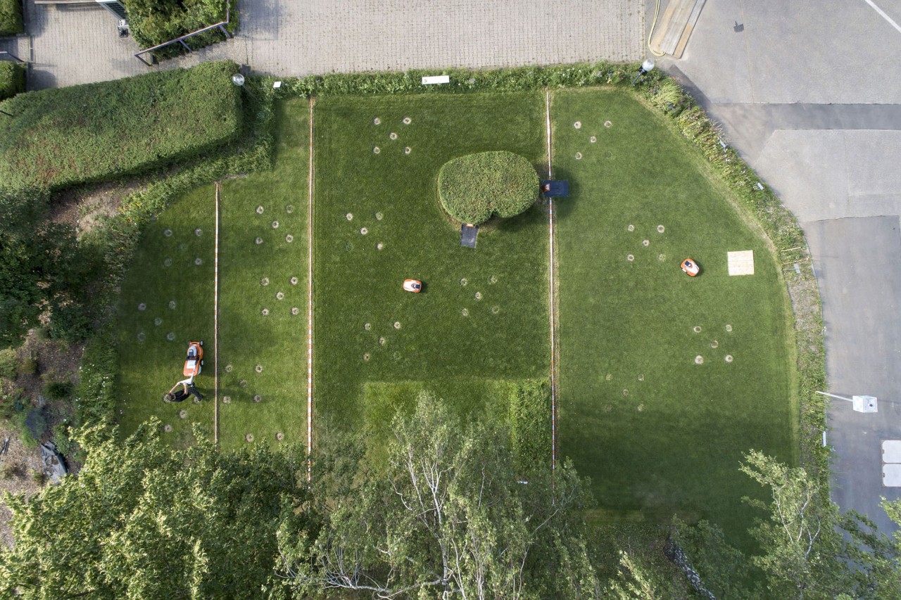 Blik van bovenaf op een in vier delen opgedeeld terrein met STIHL robotmaaiers en een grasmaaier op het bedrijfsterrein