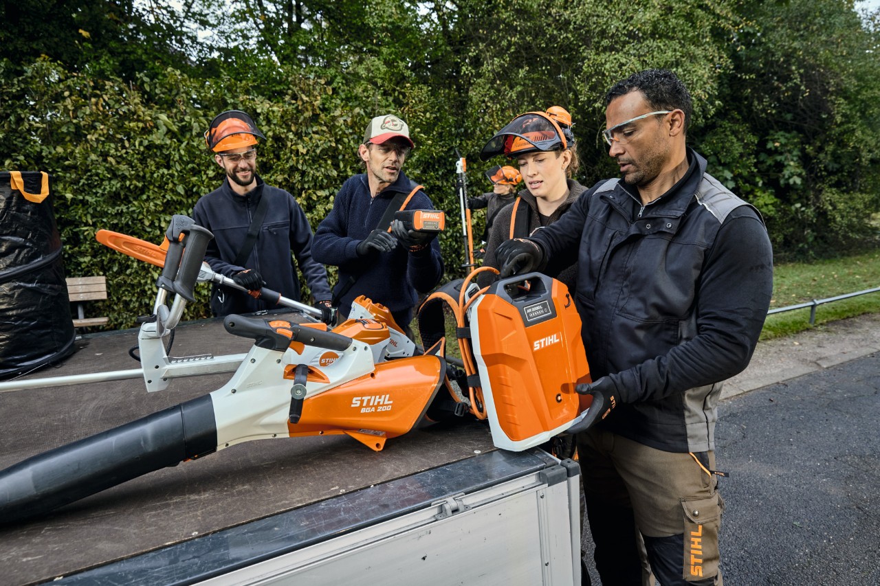 Drie mannen en een vrouw in STIHL veiligheidsuitrusting staan op de laadvloer van een voertuig met STIHL machines, waaronder de accubladblazer STIHL BGA 200 en de op de rug gedragen accu STIHL AR 2000 L 
