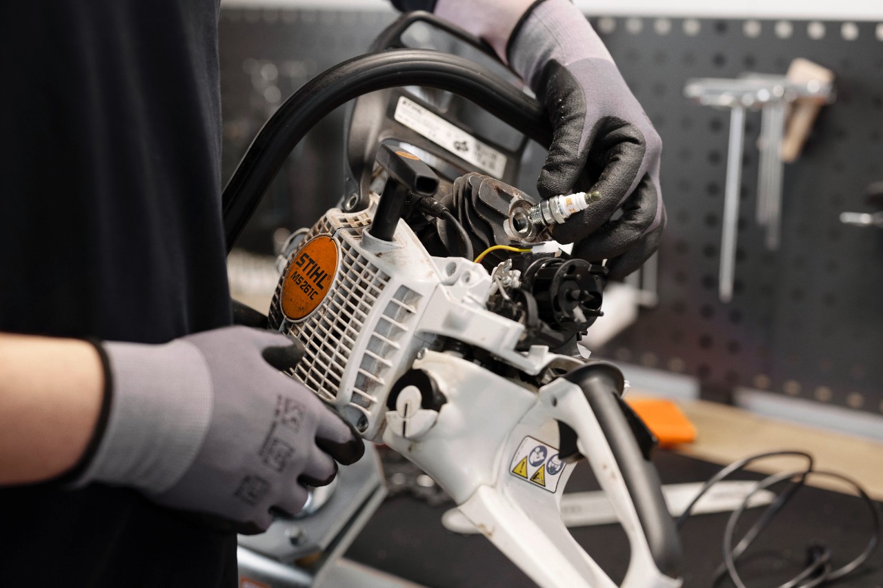 In een werkplaats staat op de voorgrond een persoon met handschoenen aan die de motor van een kettingzaag vasthoudt en een bougie verwisselt.