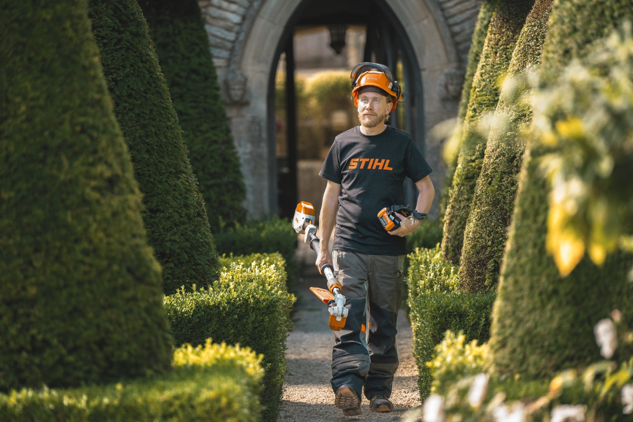 Hoofdtuinier Andrew Wain loopt met een veiligheidshelm, een STIHL T-shirt en een STIHL accumachine met een aparte accu in de hand door een tuinpad