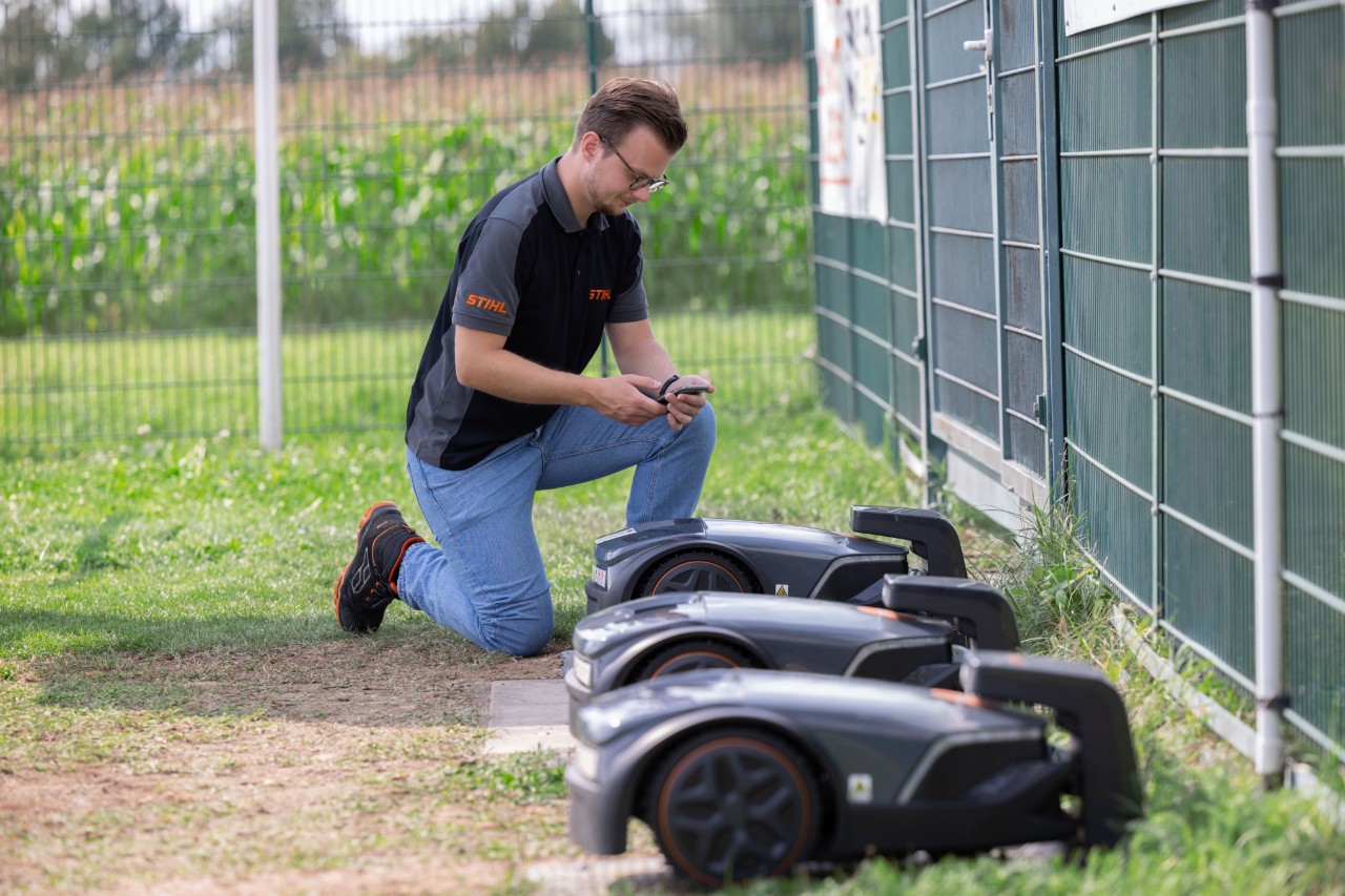 STIHL iMOW® productmanager Thilo Ehrentreich knielt naast drie robotmaaiers met een mobiele telefoon in de hand