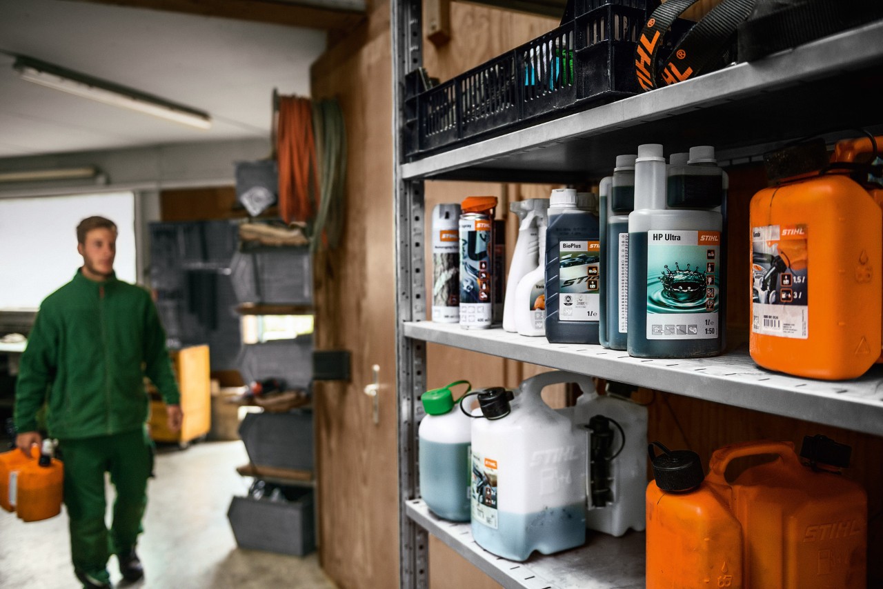Verschillende STIHL bedrijfsstoffen op een rek, op de achtergrond een man met jerrycan voor het transport van gevaarlijke stoffen 