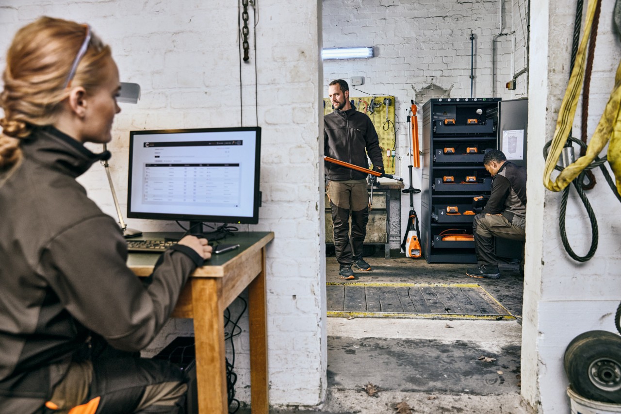 Vrouw achter een laptop in een werkplaats, rechtsachter op de foto twee mannen bij een accu-oplaadkast.