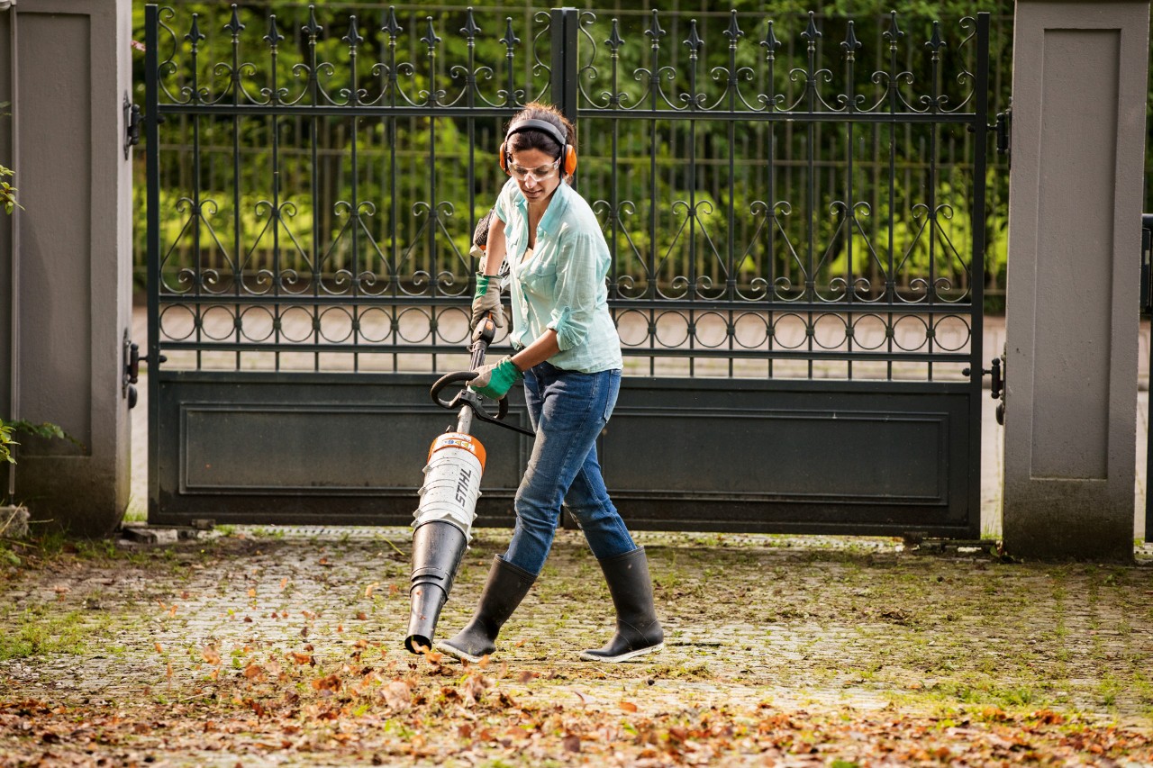 Vrouw met gezichts- en gehoorbescherming verwijdert bladeren met de STIHL combimotor KM 131 R met bladblazeropzetstuk.