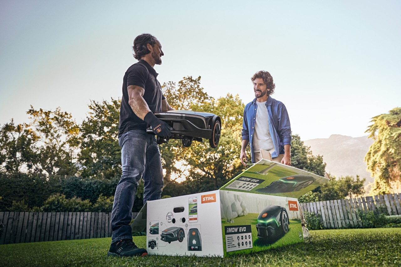 Een STIHL dealer staat met een iMOW® EVO in de hand en de verpakking van de iMOW® voor zich in de tuin van een klant om te helpen bij de installatie