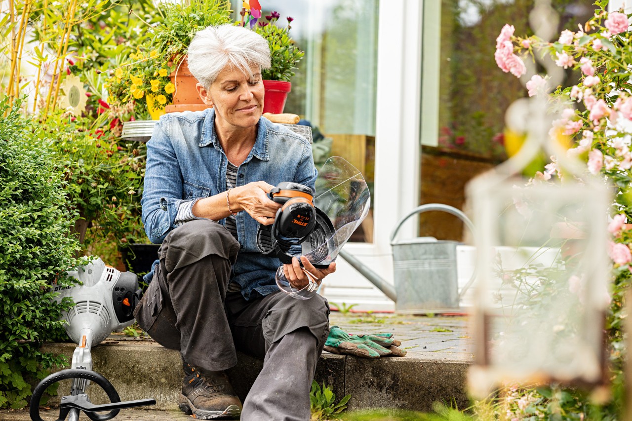 Vrouw zit tussen bloeiende planten naast een STIHL bosmaaier FS 38 en bekijkt de beschermende uitrusting