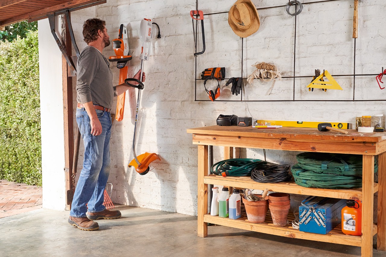 Een man in een garage neemt een STIHL elektrische bosmaaier van de muur met daarnaast een STIHL accuheggenschaar 