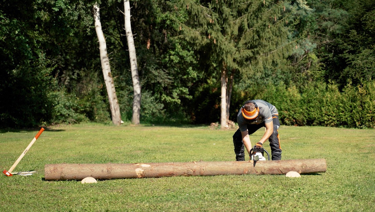 Een persoon in veiligheidsuitrusting zet een STIHL kettingzaag aan op een boomstam om brandhout te zagen.
