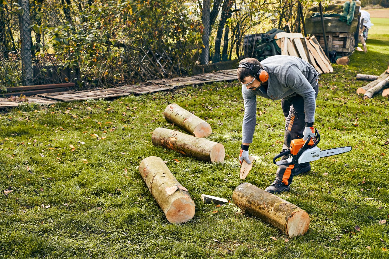 A metre-long log on grass is secured with wedges against rolling away, by someone wearing protective gear and holding a STIHL MS 180 petrol-driven chainsaw.