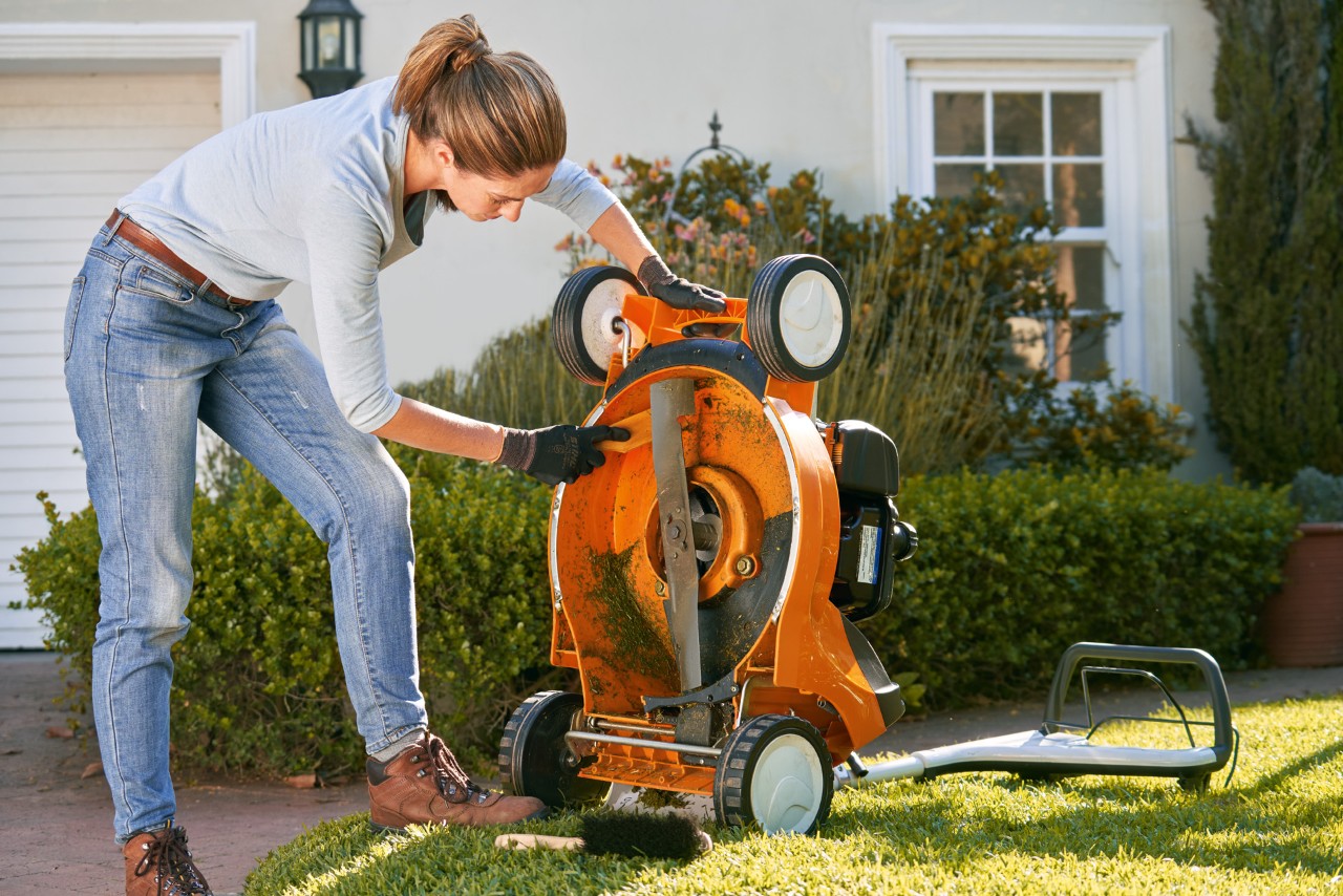 Een vrouw maakt de onderkant van een STIHL RM 448 T-C benzinegrasmaaier schoon met een spatel