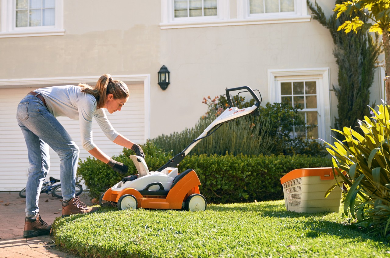 Een vrouw opent het deksel van een STIHL RMA 339 C accugrasmaaier voor een wit huis
