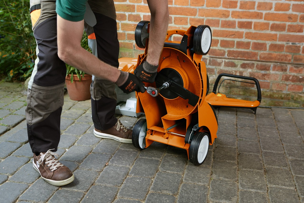 Een persoon die veiligheidshandschoenen draagt verwijdert het maaimes van een STIHL benzinegrasmaaier die op zijn kant ligt.