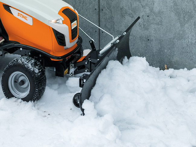Met een sneeuwploeg gemonteerd op een zitmaaier word sneeuw van voor een muur geruimd