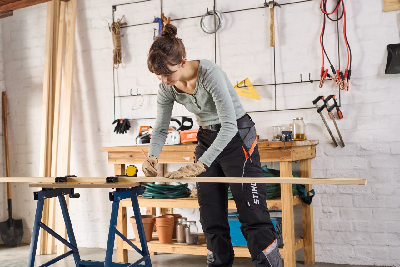 Een vrouw in een werkplaats markeert met een potlood een houten lat voor een zelfgemaakte afdekking voor een verhoogd bed