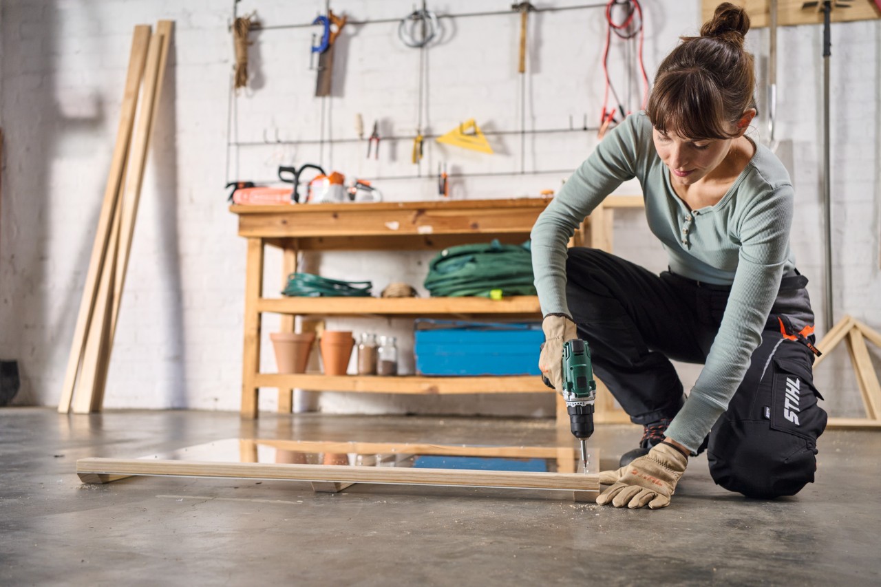 Een vrouw in een werkplaats schroeft een houten lat vast aan een plexiglas plaat