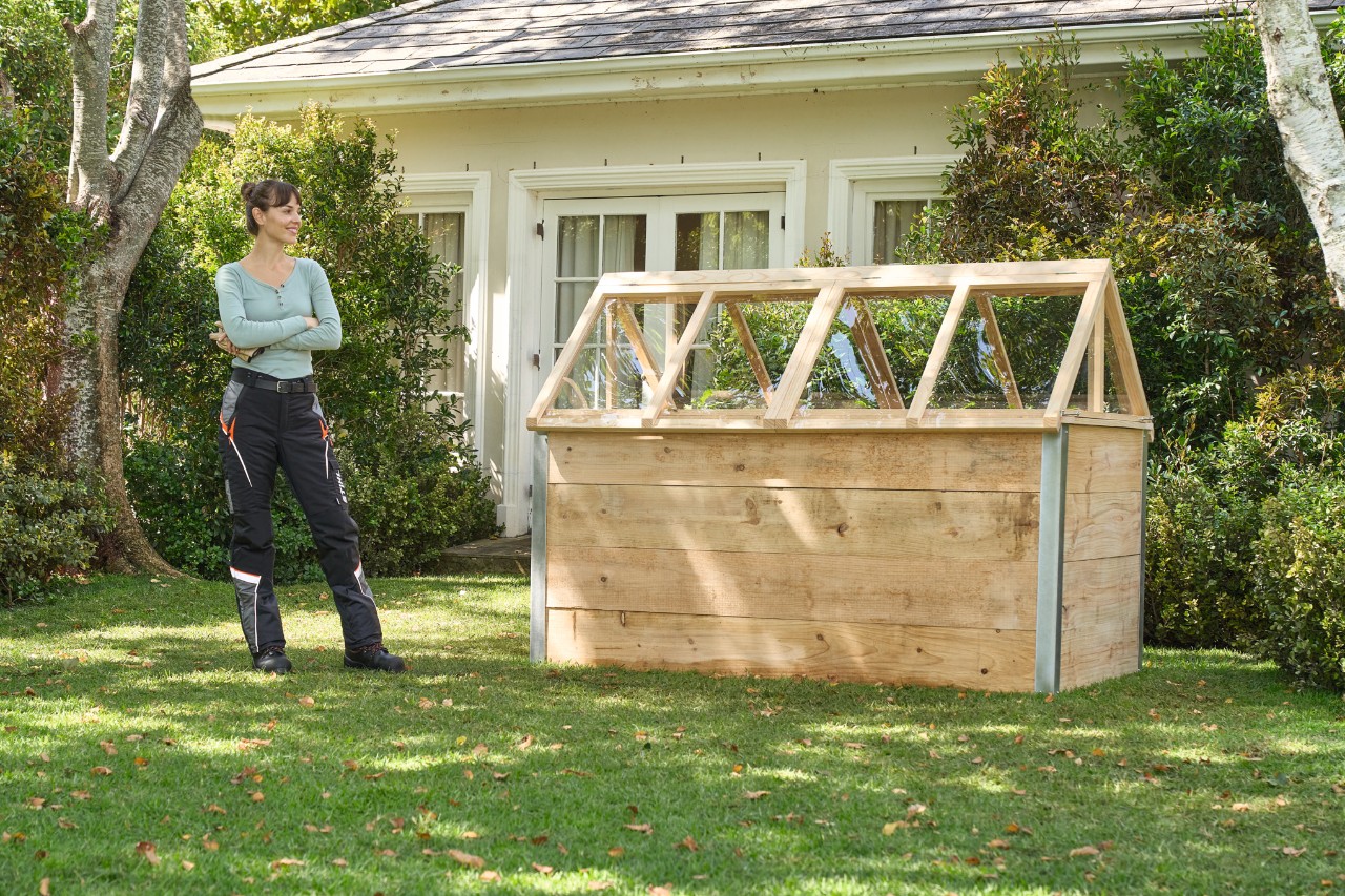 Een vrouw op een tuinperceel staat tevreden en lachend met gekruiste armen voor een verhoogd bed met een zelfgemaakte afdekking 