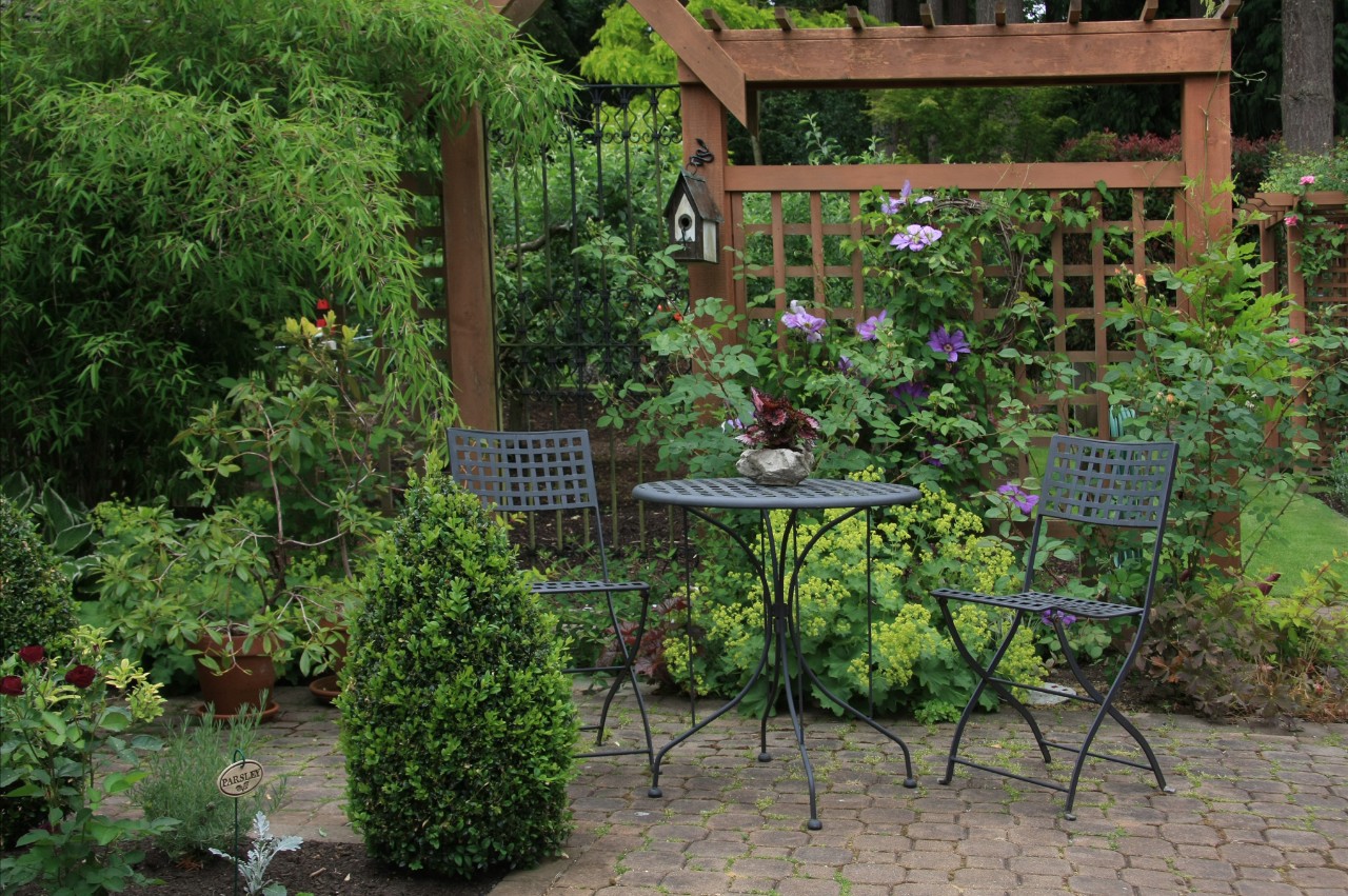 Begroeid houten leirek tussen groene struiken met ervoor een tuintafel en twee stoelen