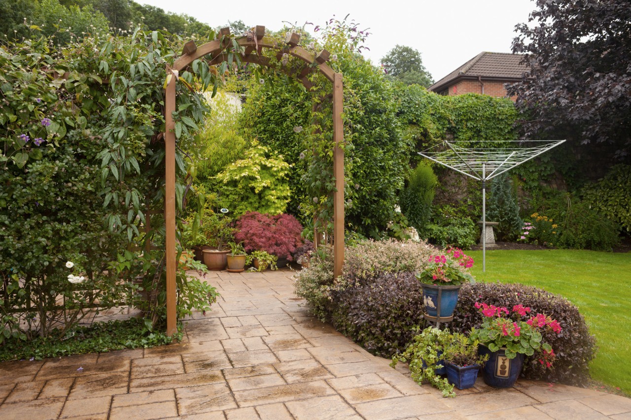 Arche pour rosiers en bois recouverte par la végétation au-dessus d’une allée de jardin, pelouse avec sèche-linge à côté
