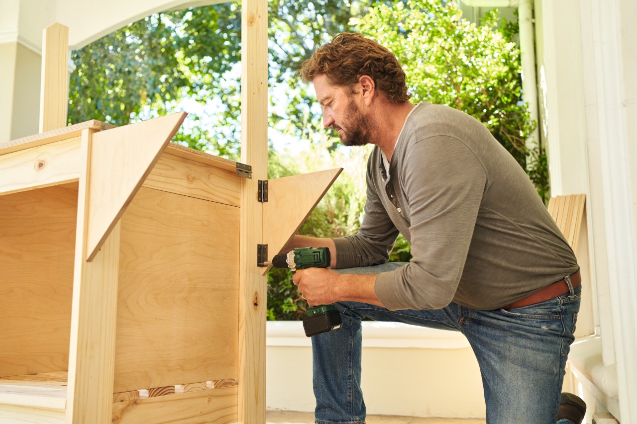 Man op een veranda schroeft scharnieren met hoekstukken vast aan een plantentafel