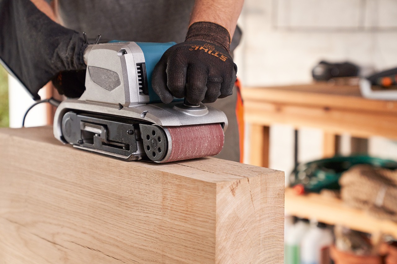 Close-up van iemand met STIHL veiligheidshandschoenen die met een schuurmachine hout schuurt in de werkplaats