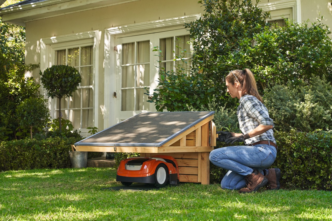 Vrouw op een tuinperceel met een garage voor een zelfgebouwde robotmaaier, inclusief een STIHL robotmaaier, met een huis op de achtergrond. 