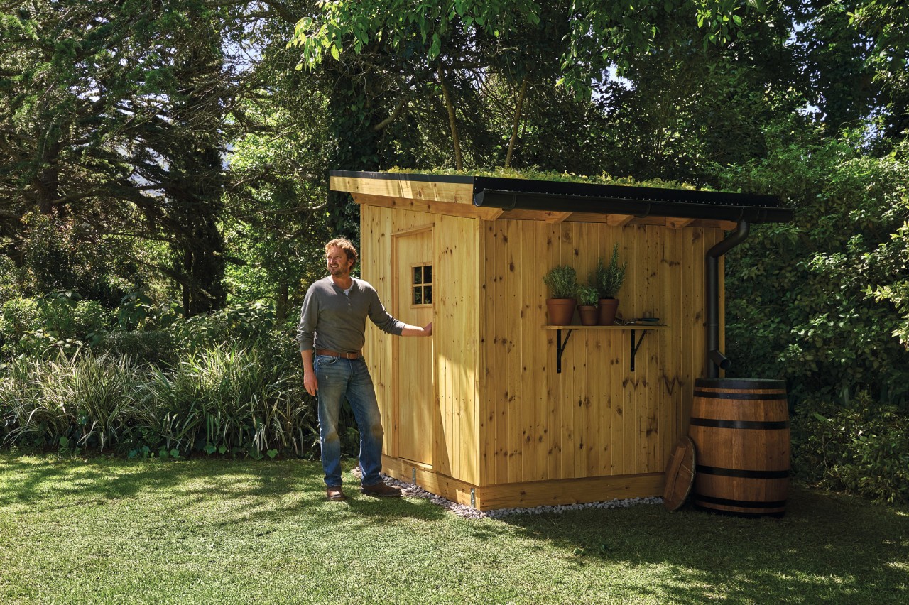 Man in spijkerbroek op een tuinperceel voor een zelfgebouwd gereedschapsschuurtje.