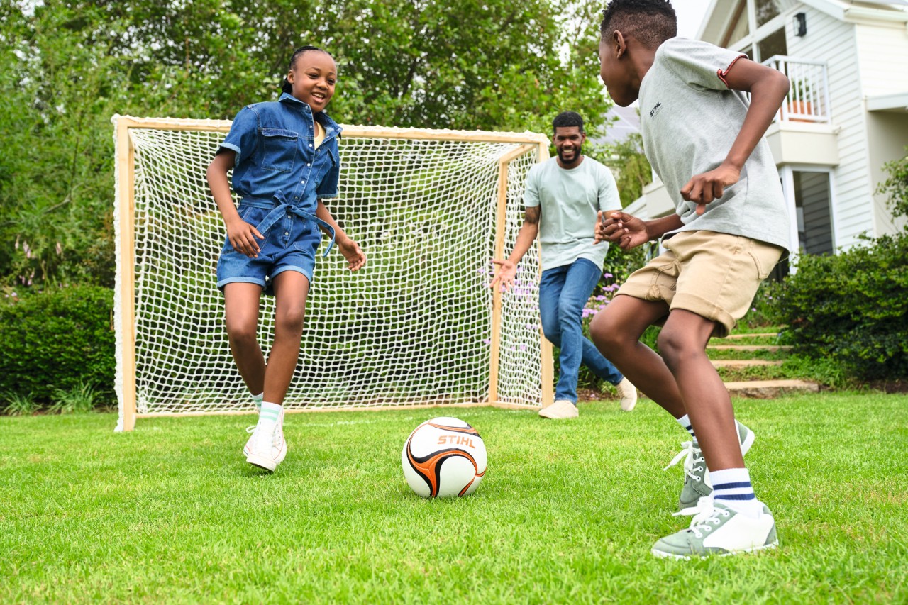 Trois enfants jouent au football dans le jardin avec un but de football en bois.