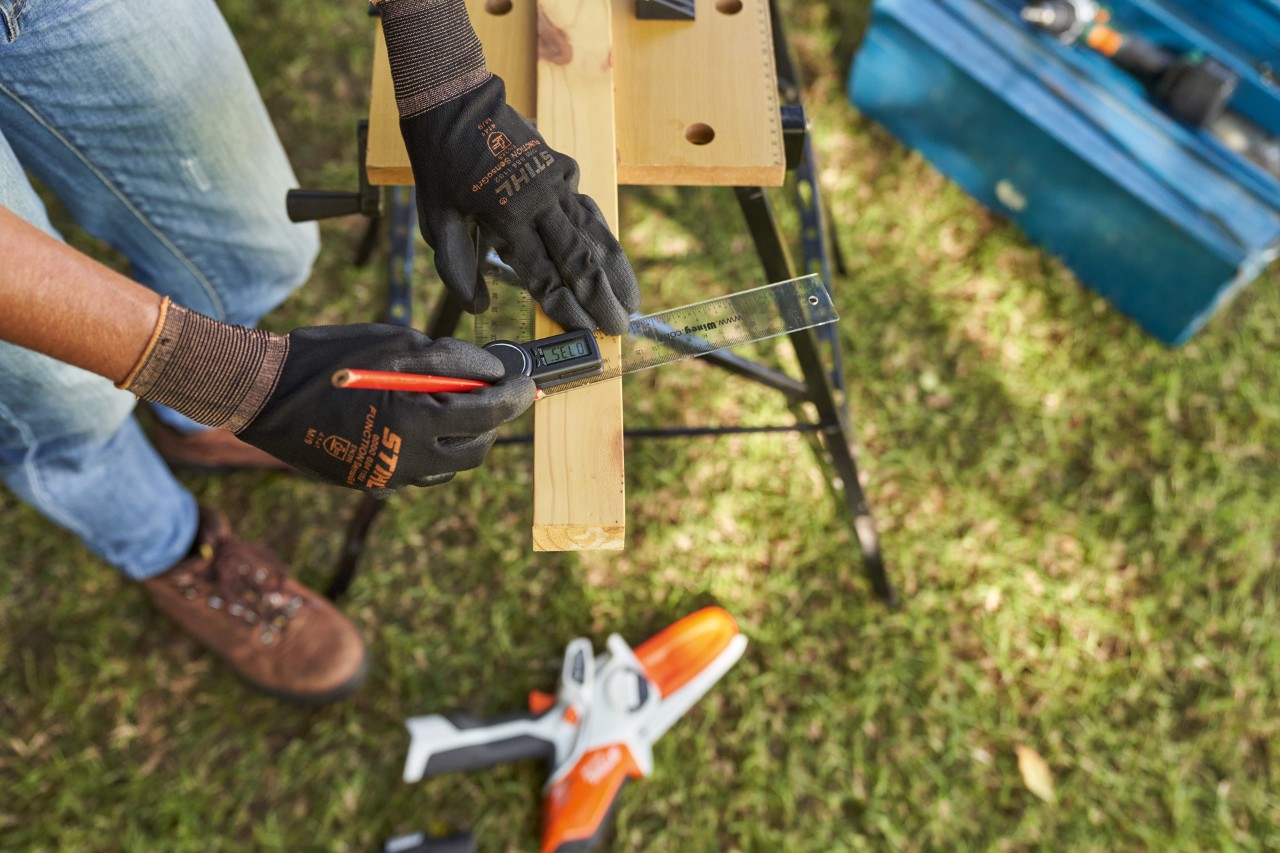 Close-up: vrouw met werkhandschoenen meet bouwhout met op de grond de STIHL GTA 26 accusnoeizaag