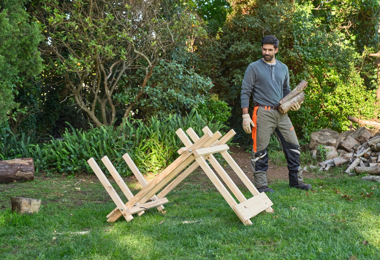 Een man in een tuin draagt houtblokken naar een zelfgemaakte zaagbok