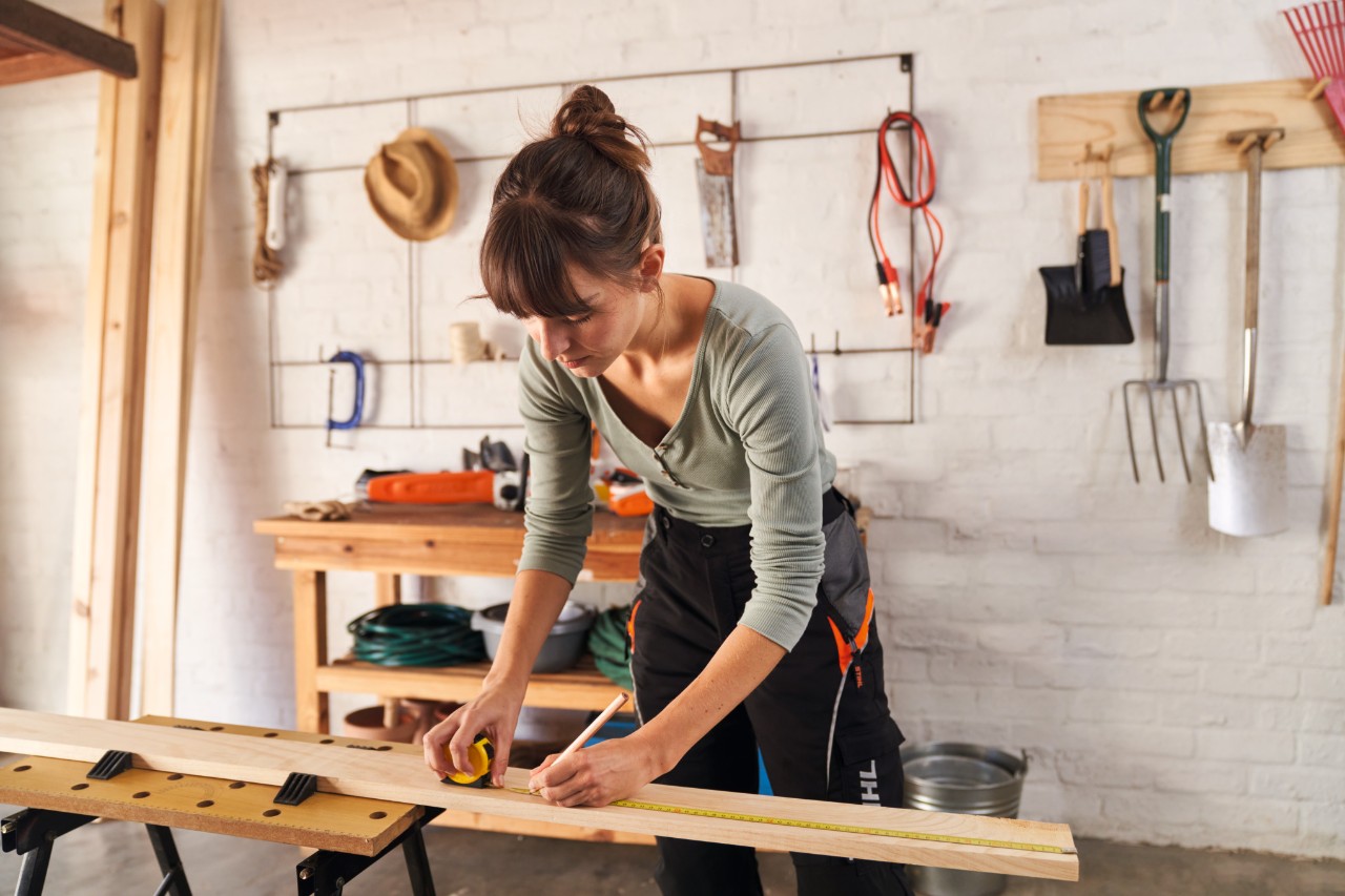 Een vrouw meet houten onderdelen op de werkbank met een meetlint en markeert de lengtes met een potlood