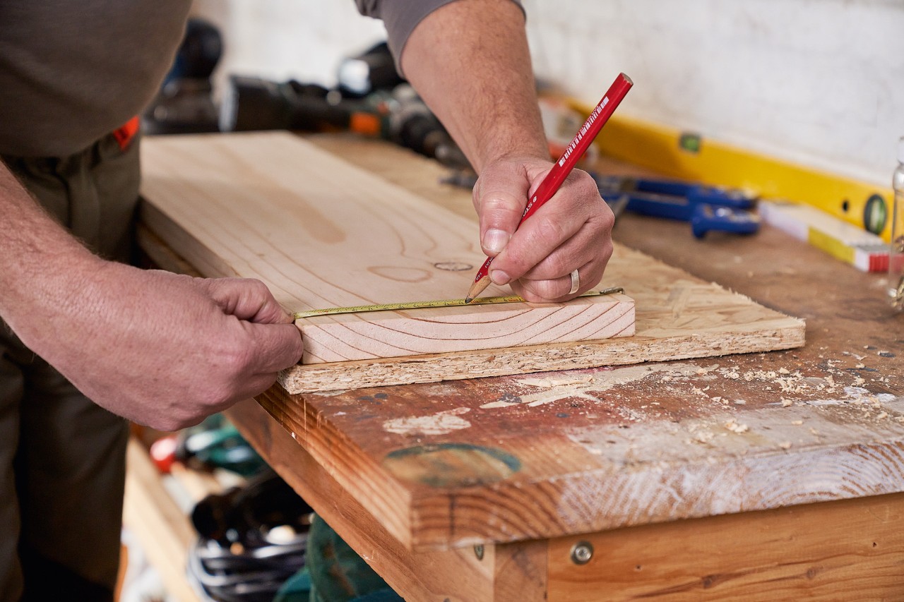 Een man markeert boorgaten op een houten plank om een eigen een tuingereedschaphouder te maken