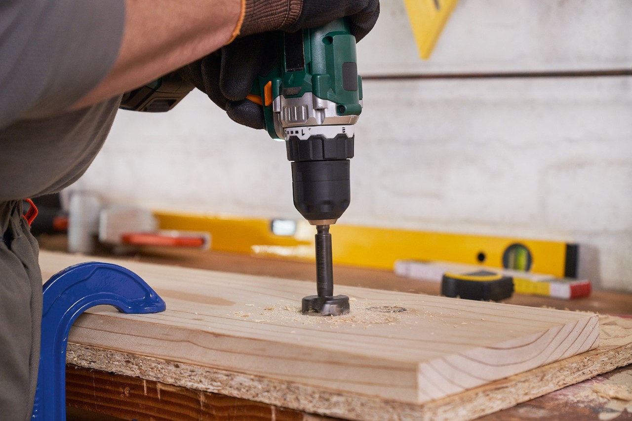 Een man boort met een speedboor gaten in een houten plank voor een tuingereedschaphouder
