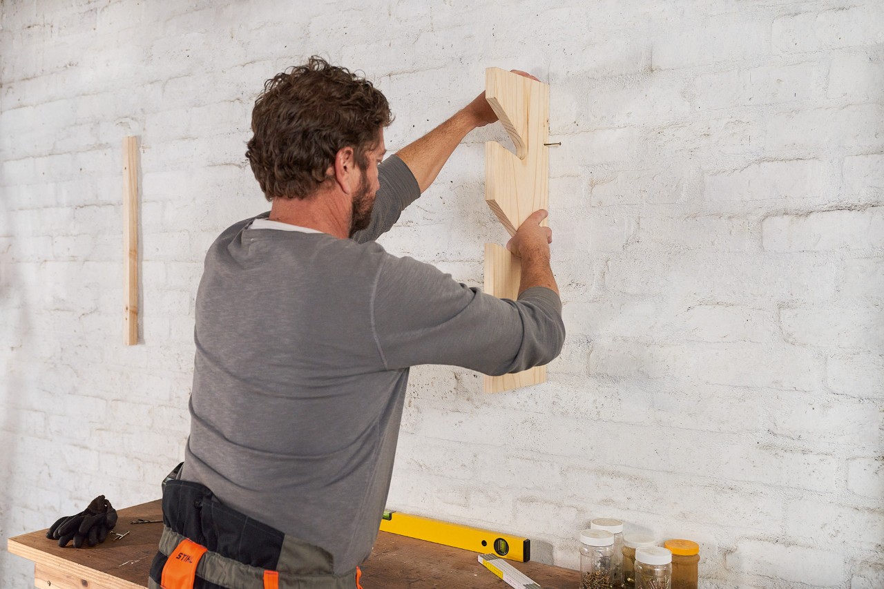 Een man brengt deuvels in een houten lat aan de muur aan om een eigen tuingereedschaphouder te maken 