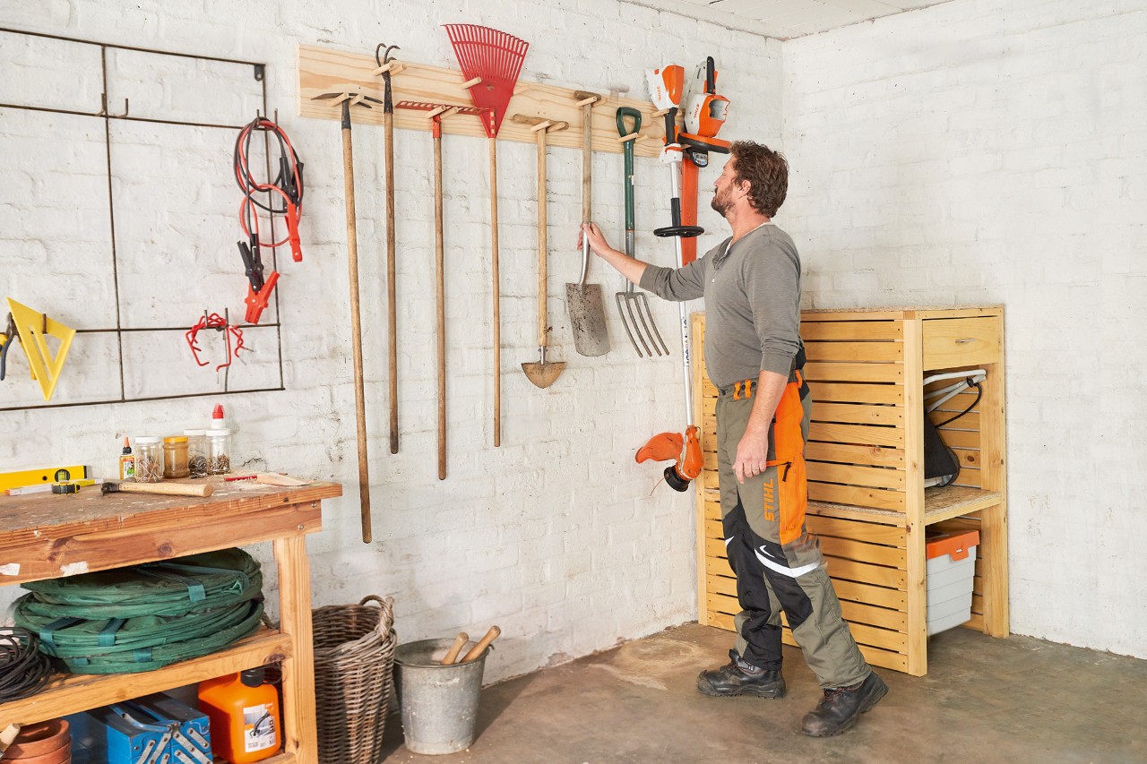 Man in STIHL werkbroek hangt een schop aan een zelfgemaakte tuingereedschaphouder naast een STIHL grastrimmer 