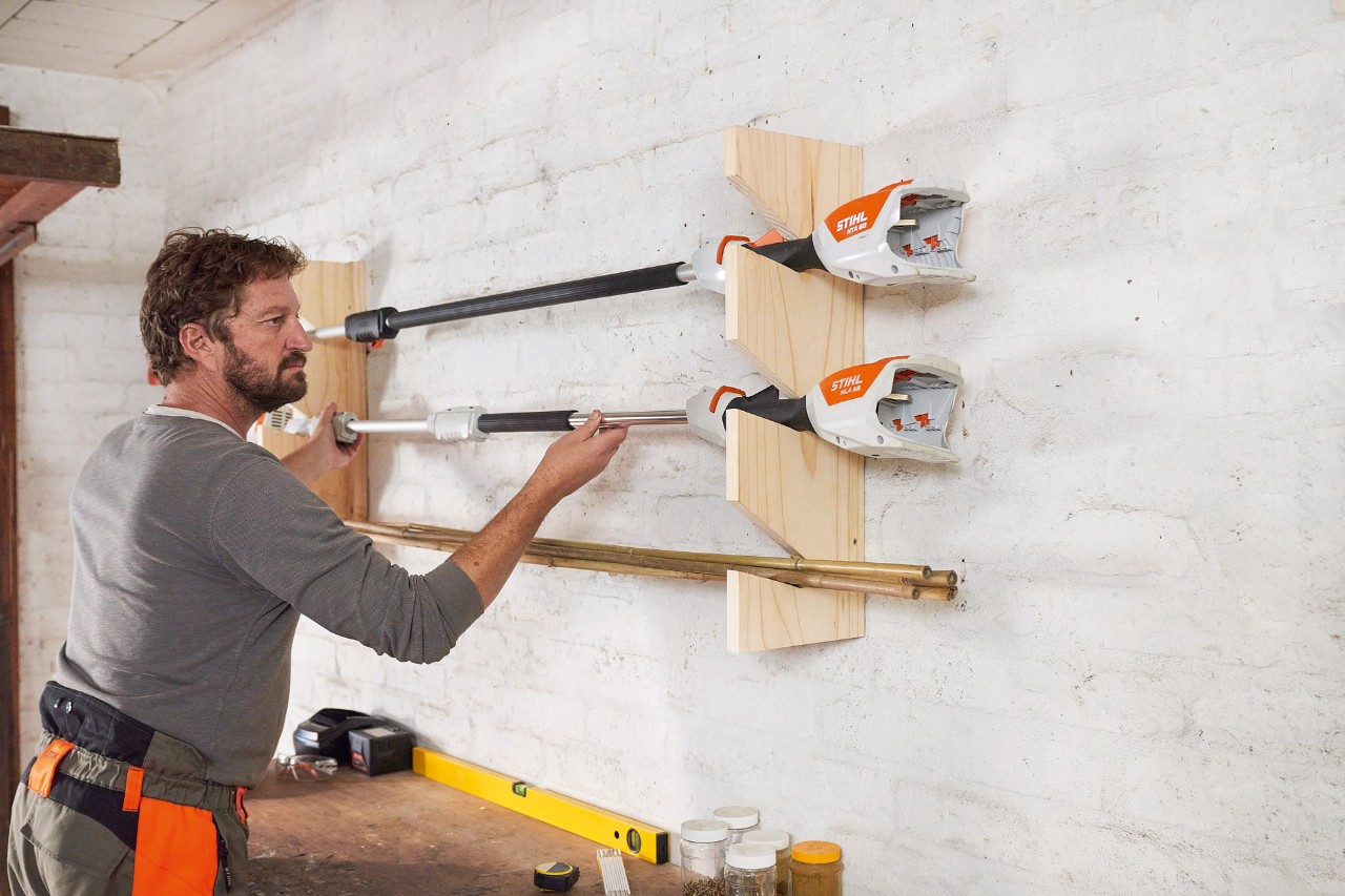 Een man hangt een STIHL grastrimmer aan een zelfgebouwde tuingereedschaphouder aan de muur