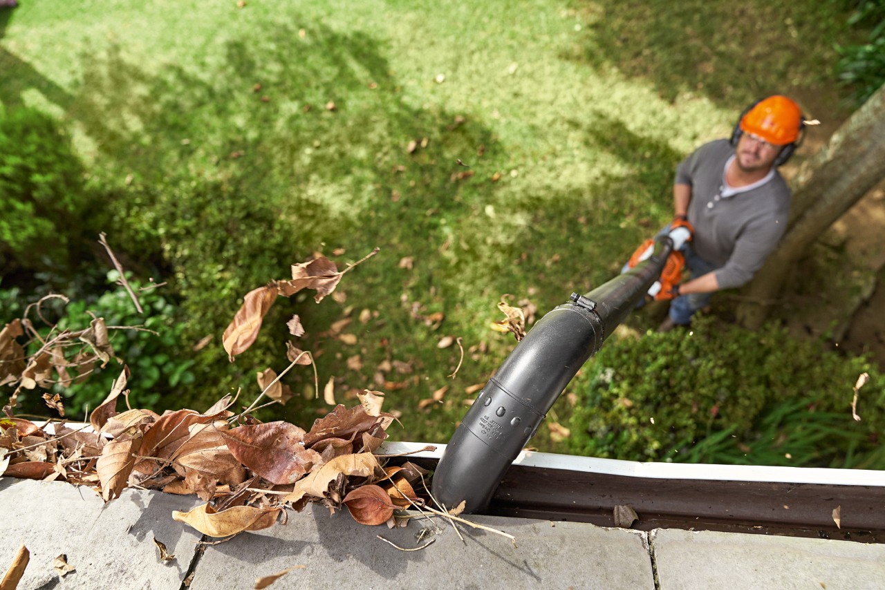 Foto in bovenaanzicht van een dakgoot vol bladeren die wordt gereinigd door een man die op de grond staat met een STIHL bladblazer en dakgootreinigingsset.