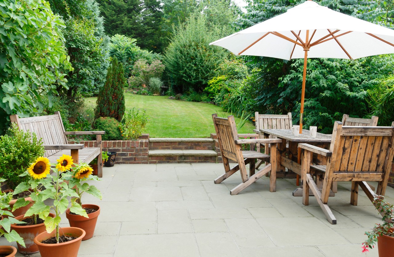 Houten meubelen op een ruim terras met betontegels in een grote groene tuin