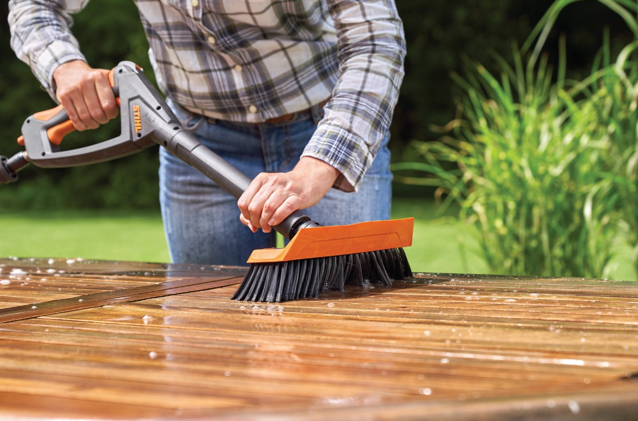 Een vrouw die een houten tuintafel schoonmaakt met een STIHL RE 130 Plus hogedrukreiniger met wasborstel