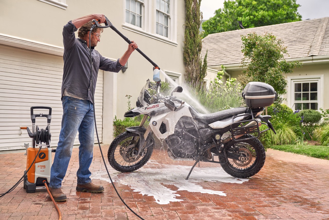 Een man gebruikt de STIHL RE 130 plus hogedrukreiniger om autoshampoo op een motor te spuiten 