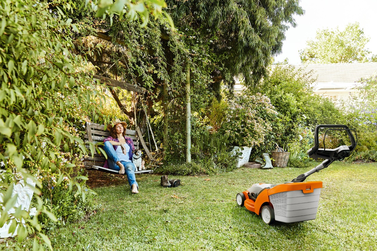 Vrouw ontspant op een tuinschommel naast een STIHL RMA 339 accugrasmaaier op een groen grondstuk