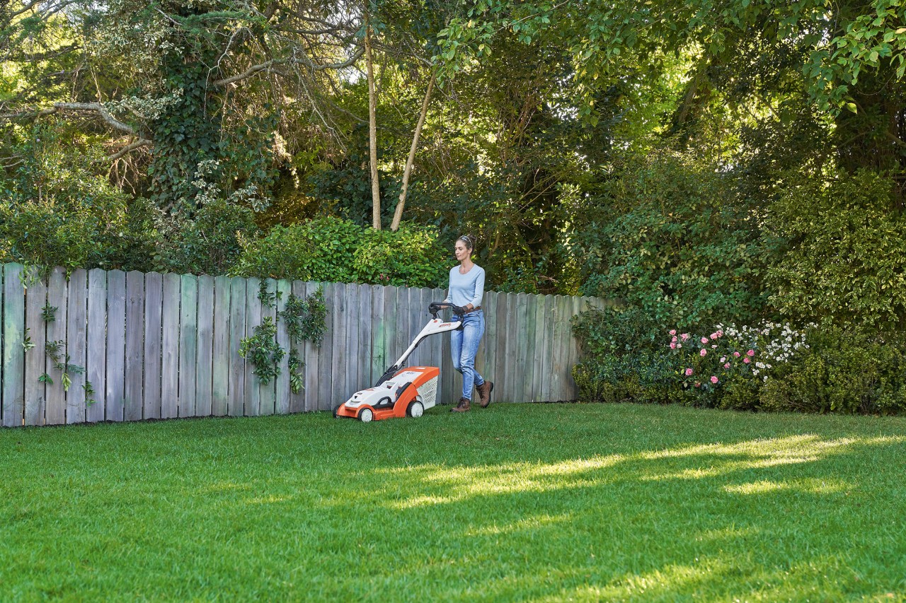Vrouw maait groene grasmat voor een houten hek met STIHL RMA 339 C accugrasmaaier.