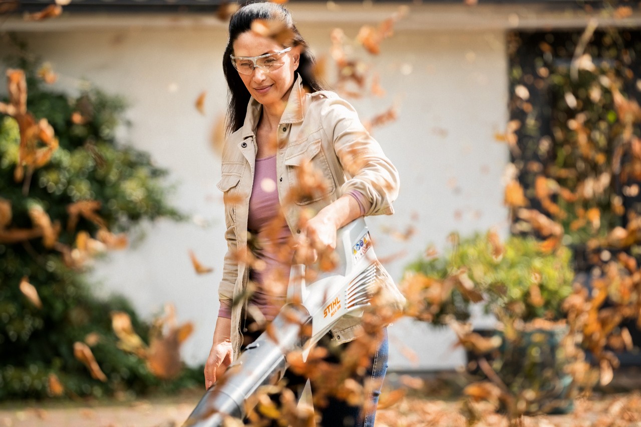 Een vrouw die een STIHL BGA 56 accubladblazer gebruikt om in het voorjaar op te ruimen, met bladeren op de voorgrond