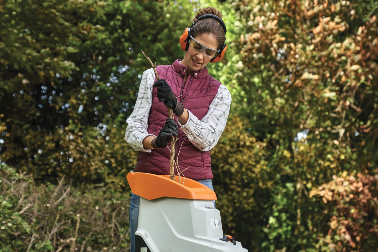 Een vrouw in beschermende werkkleding hakselt snoeisel van een thujaheg, op de achtergrond bomen