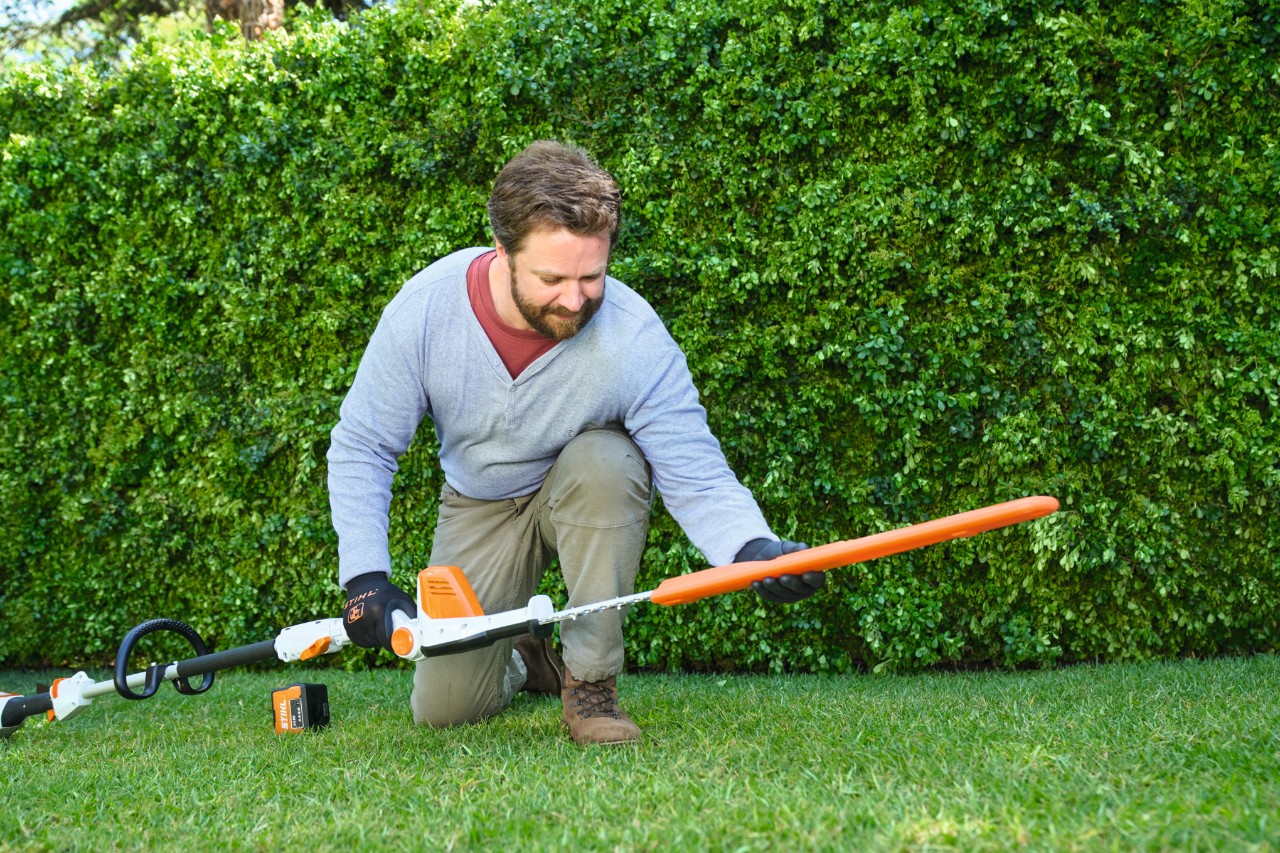 Een man staat voor een heg en legt een STIHL HLA 56 accuheggensnoeier op het gazon neer