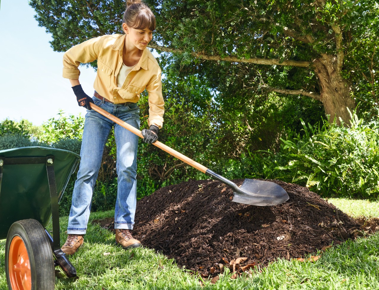 Een vrouw stort met de spade een laag grove compost op het heuvelbed