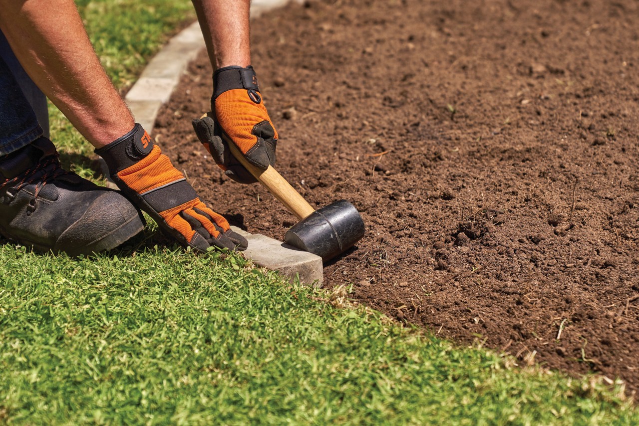 Een persoon die handschoenen draagt met een hamer om een rand met stenen rond een tuinbed te maken