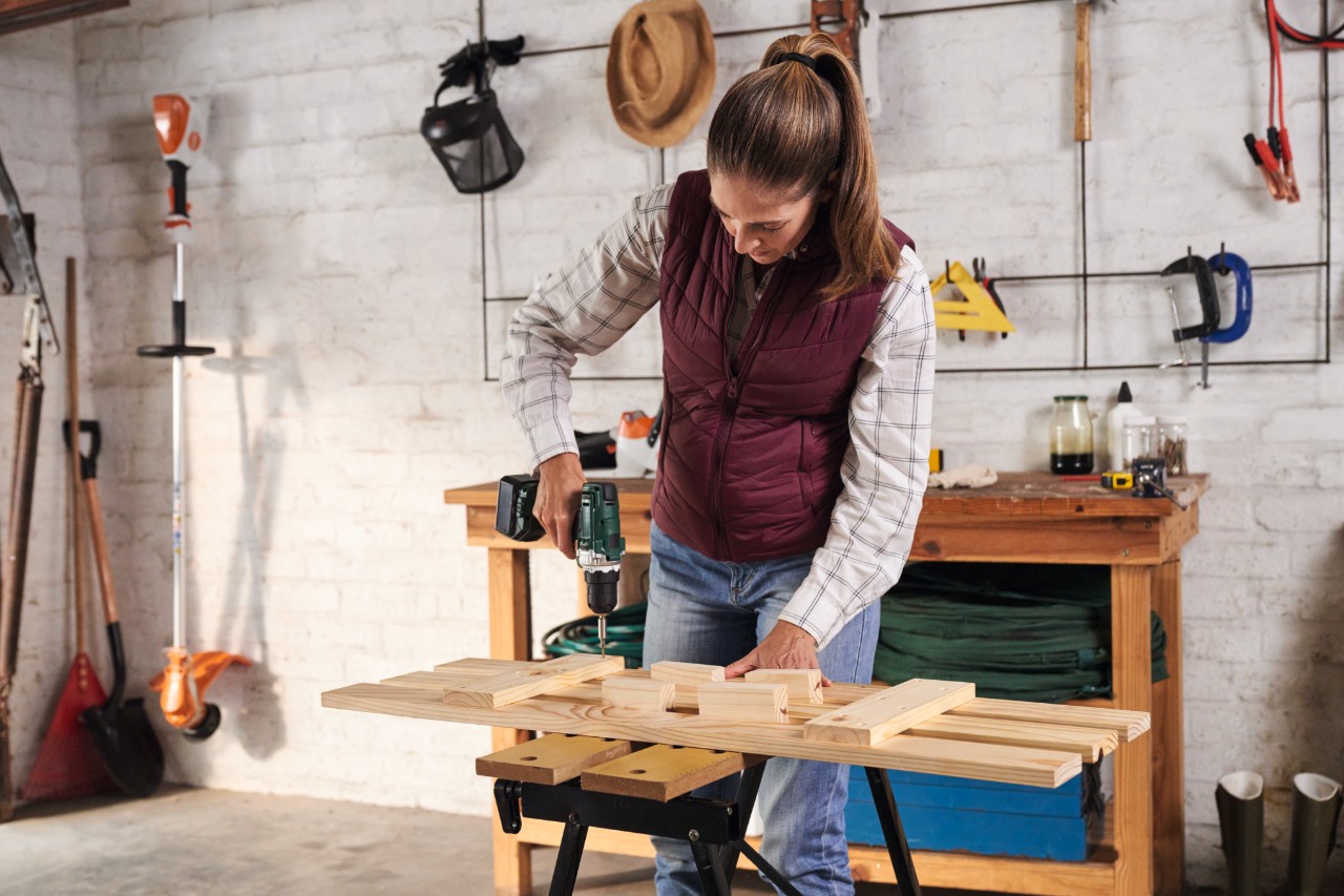 Vrouw met geruit hemd schroeft latten aan dwarsbalken vast op een werktafel, met op de achtergrond een gereedschapswand