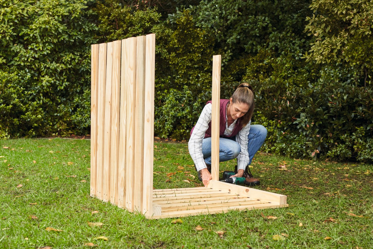 Vrouw in jeans bevestigt de latten voor de zijwanden aan de compostbak met een accuschroevendraaier in een tuin
