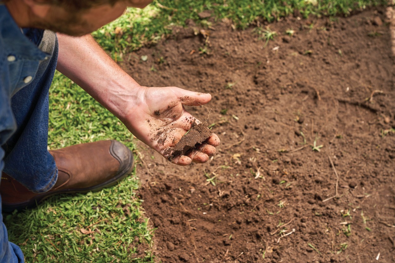 Man houdt een stuk aarde in z'n hand, op de achtergrond bruine aarde en gras